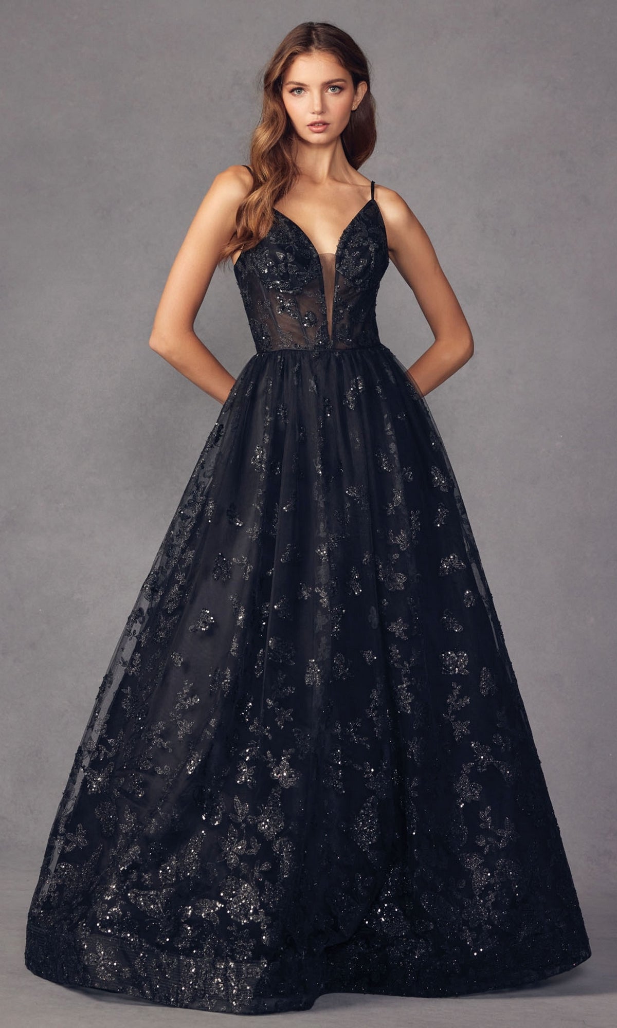 A woman stands against a neutral gray background wearing the Sheer-Corset Glitter-Print Long Prom Dress 2413—a sleeveless, floor-length black A-line gown with a deep V neckline and shimmering floral patterns.